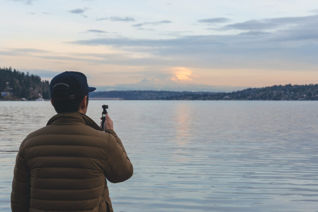 image of guy in green jacket and blue hat using the dji pocket 2 camera in front of a body of water