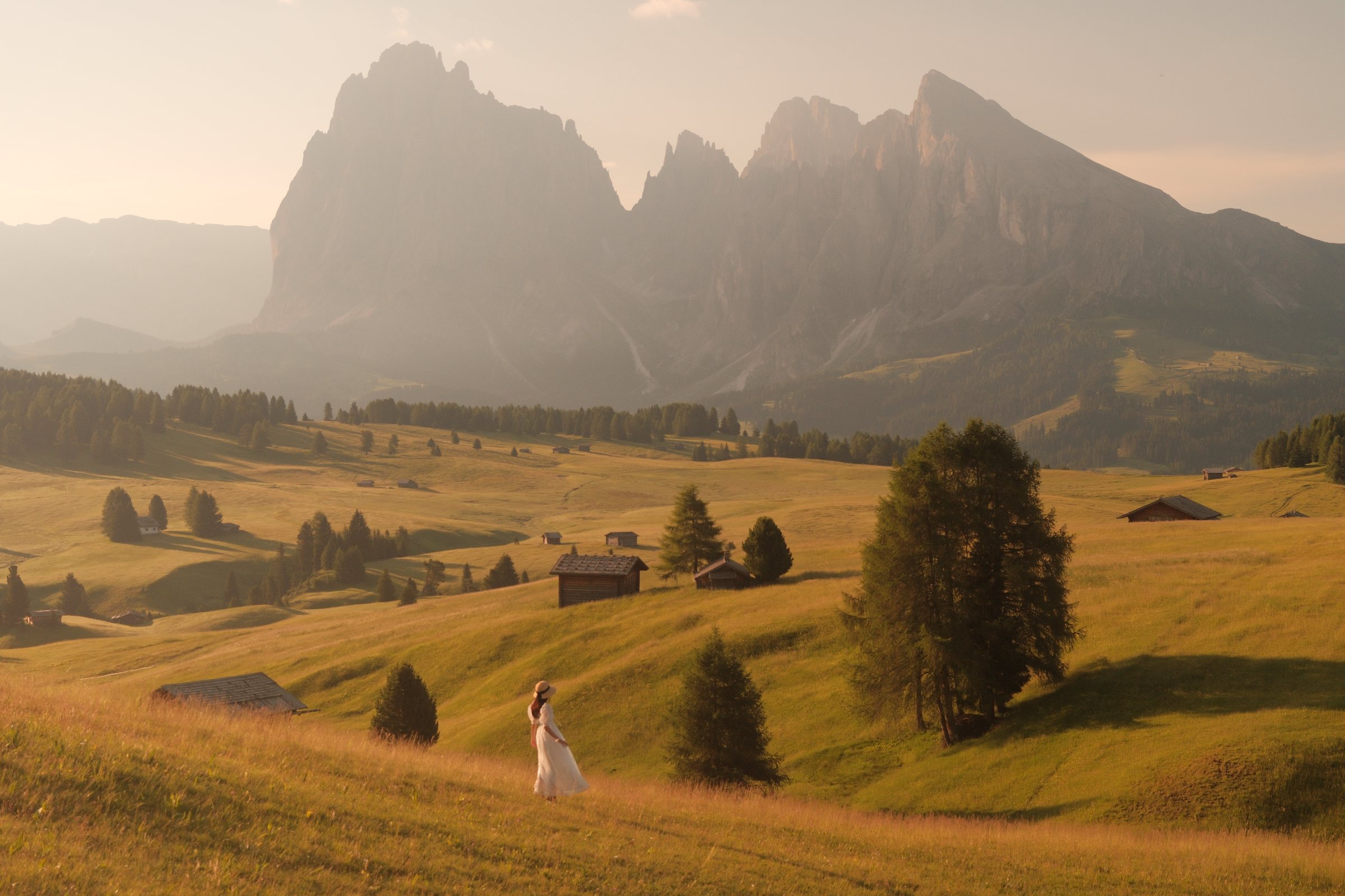 A woman in a white dress walks through golden meadows beneath the Dolomites at sunrise