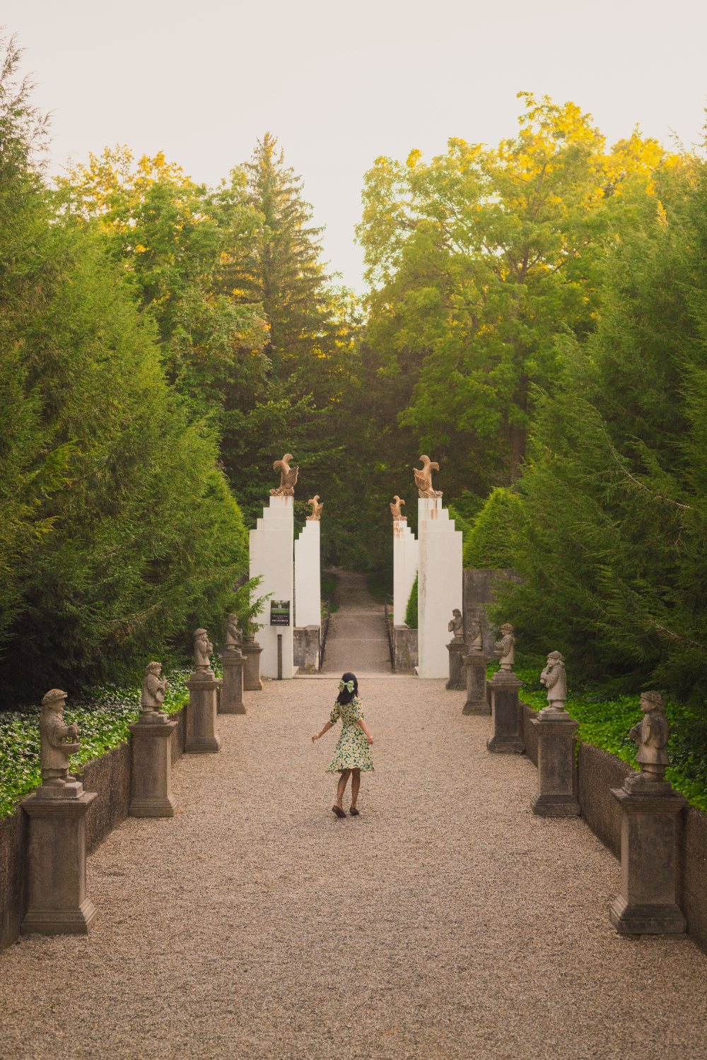 A woman twirls on a gravel path lined with statues at Allerton Park